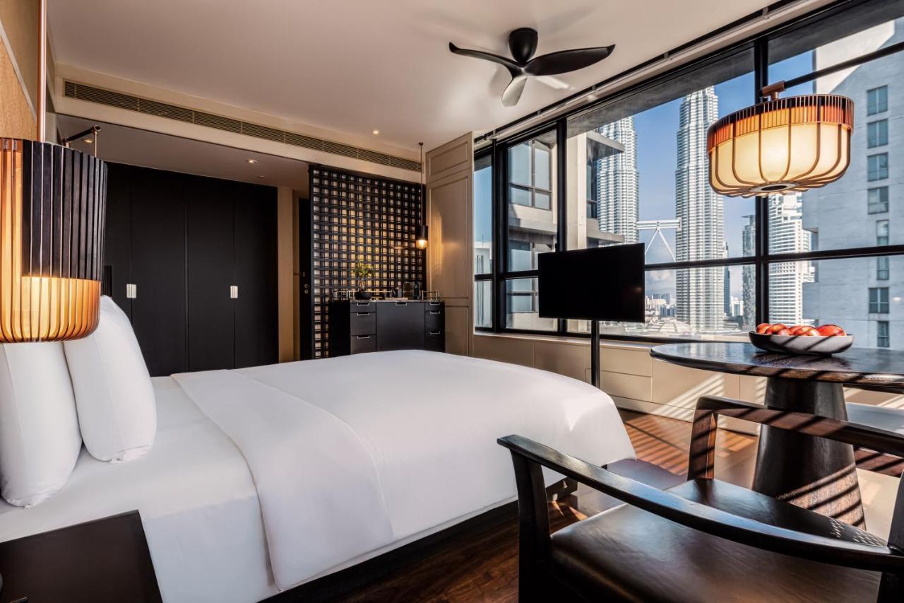 The Ruma Hotel And Residences Kuala Lumpur Exteriér fotografie The photo shows a modern hotel room featuring a large bed with white linens. There's a ceiling fan above and large windows letting in natural light, providing a view of a cityscape outside. To one side, there's a small seating area with a round table